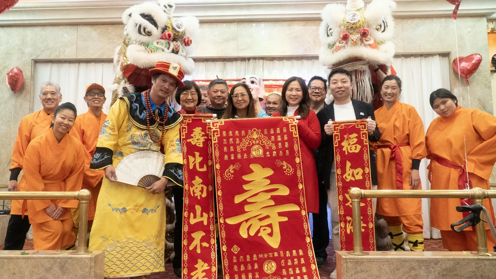 Group photo at a festival celebration: performers and guests with lion dancers and a traditional longevity scroll