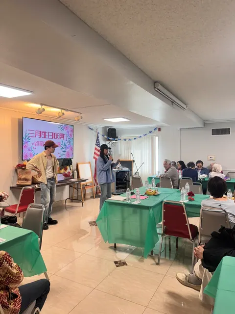 Hosts opening the March birthday celebration in front of the screen as residents sit at green tables
