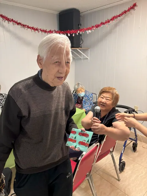 A resident happily receives a gift at the birthday party while another resident offers wishes on the microphone
