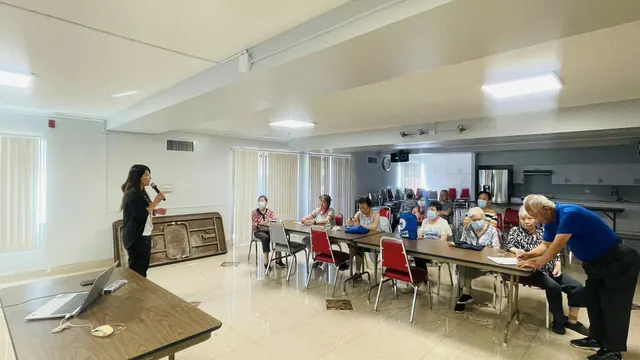 The presenter holds a microphone while engaging with seated seniors